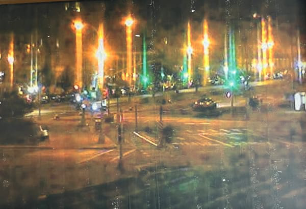Nighttime city intersection with cars, traffic lights, and tall streaks of reflected light.