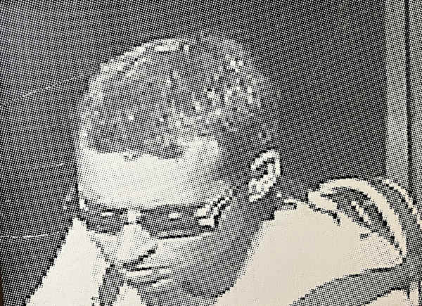 Black and white halftone portrait of a seated man wearing glasses with one hand raised.